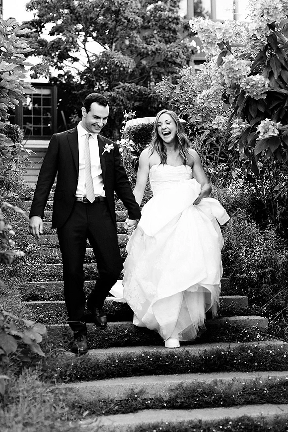 Couple portrait in a black and white wedding portrait as bride in strapless gown and veil laughs holding hands with groom on garden steps