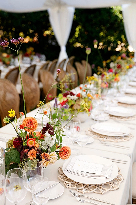 Reception tablescape with wildflower wedding centerpiece featuring orange dahlias, woven placemats, rattan chairs under a draped tent canopy
