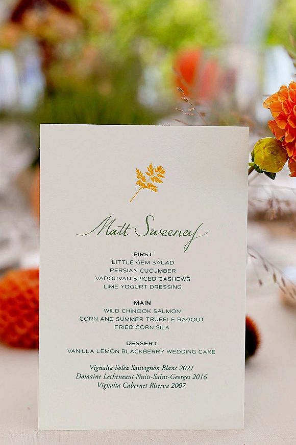 Wedding menu card with calligraphy wedding menu text and leaf illustration, accented by orange roses on a table with blurred greenery
