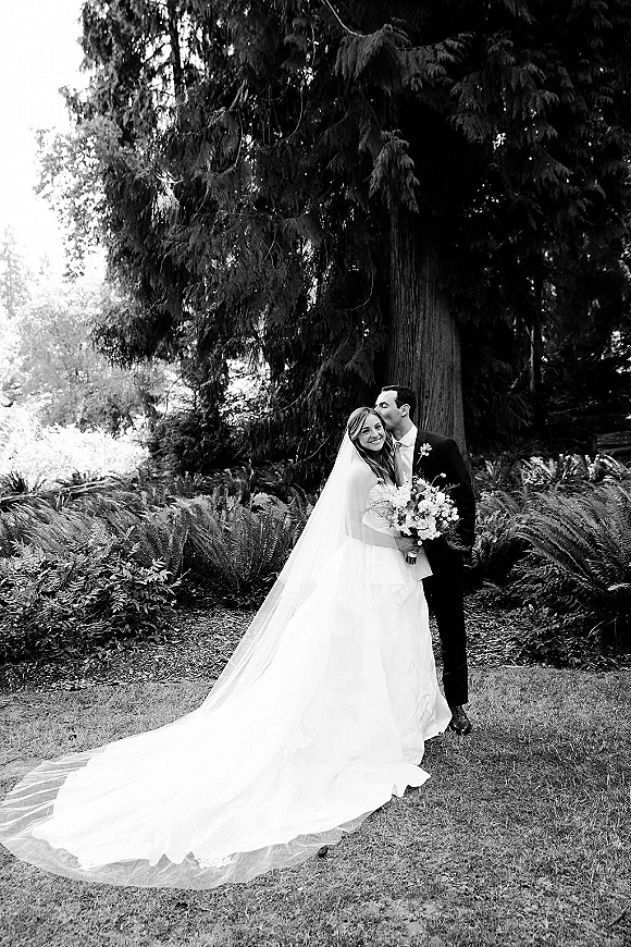 Couple portrait in a black and white wedding portrait style, groom kisses bride’s forehead as she holds a bouquet in a forest of tall trees