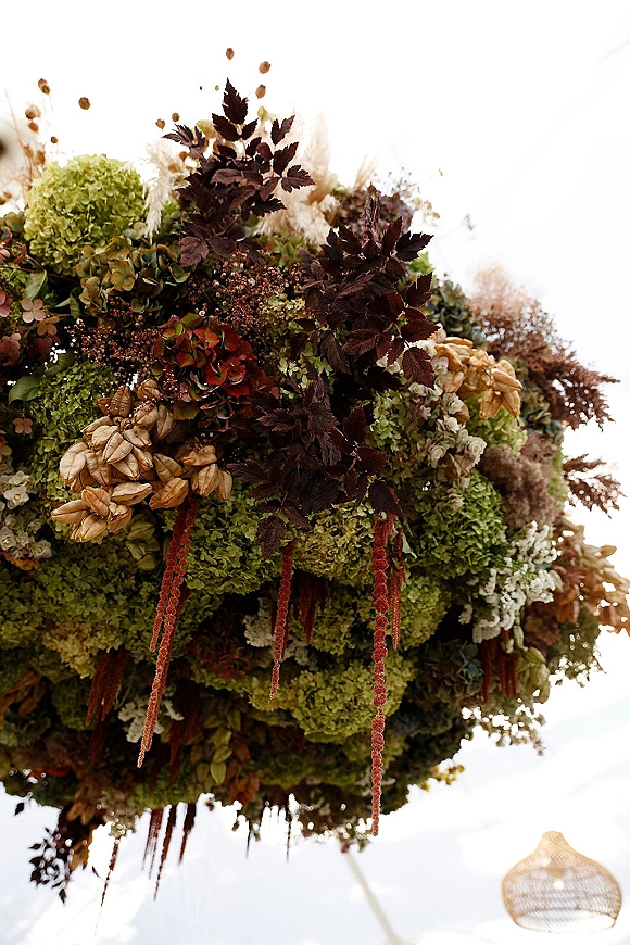 Hanging floral installation with cascading greenery and hydrangea blooms, moody burgundy foliage and dried florals under a white tent ceiling