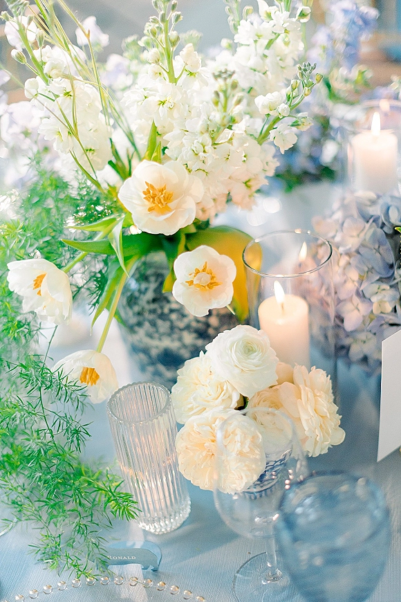 Wedding centerpieces with a white floral centerpiece in blue and white vases, greenery, and pillar candles on linen table with bokeh lights