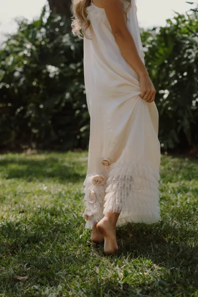 Wedding dress detail with ruffle hem wedding dress lace trim and rosette appliques as barefoot bride walks on a sunlit lawn with trees