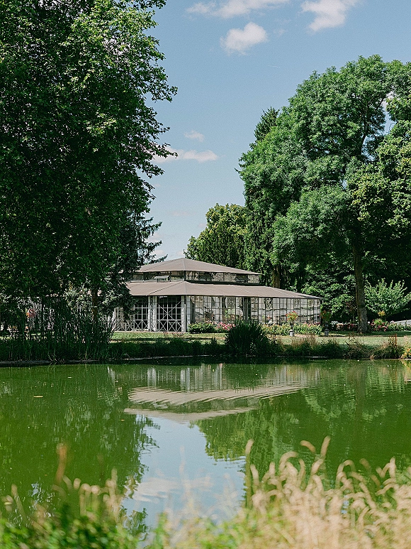 Wedding venue exterior with a glasshouse greenhouse and flower beds beside a pond, framed by trees, lawn, and cloudy sky