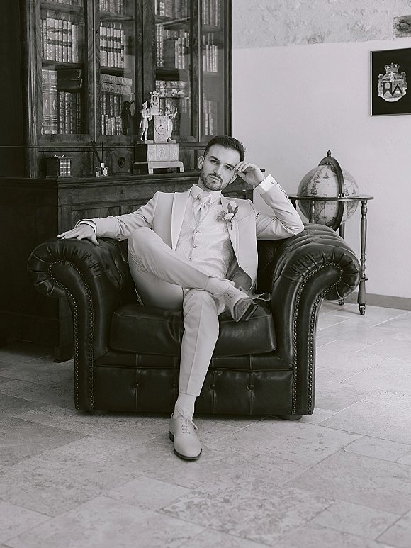 Groom portrait in a light beige suit seated in a leather armchair, tie and boutonniere visible, with a bookcase and globe bar behind