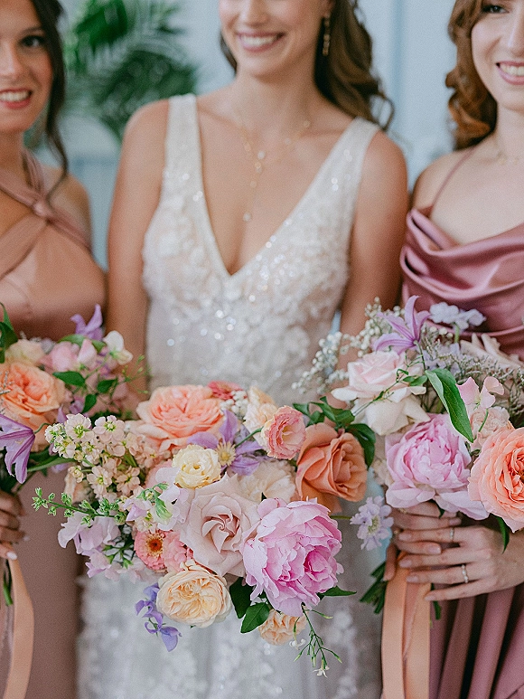 Bridesmaid bouquets of pink and peach bouquet roses and peonies held by women in blush and mauve dresses indoors by a potted plant
