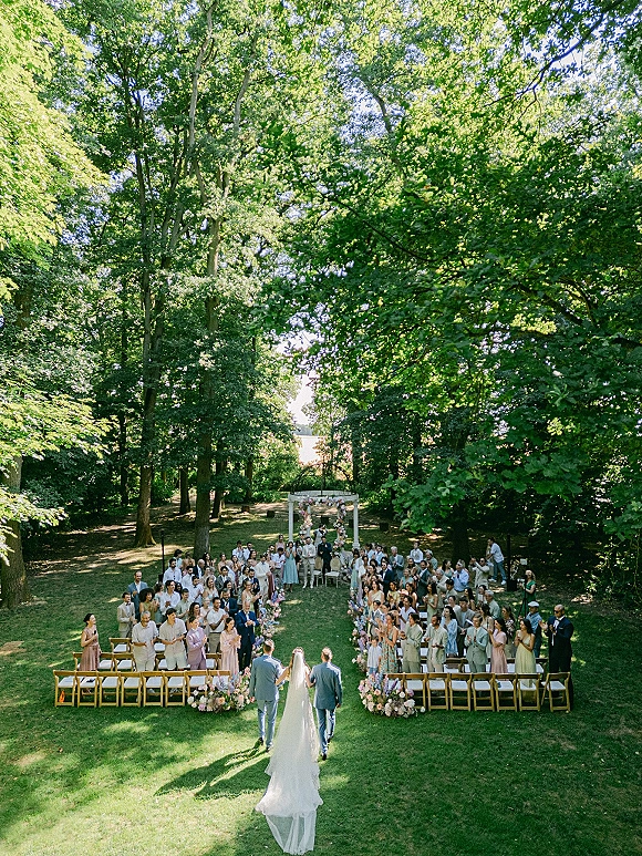 Wedding ceremony in a forest clearing with guests standing beside a floral-lined aisle leading to a circular wedding arch under sunlight
