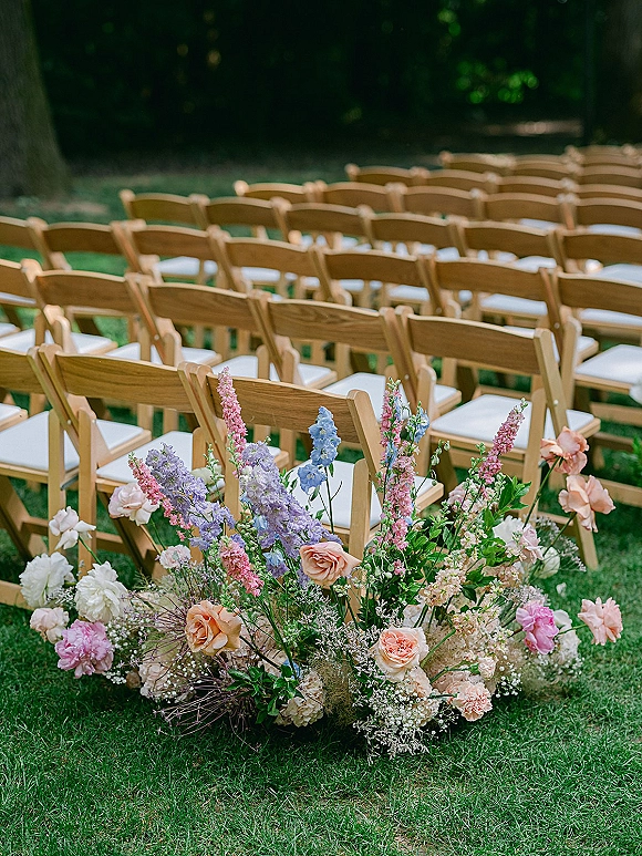 Ceremony seating with outdoor ceremony chairs, wood folding chairs and white cushions beside a meadow flower ground arrangement on a garden lawn