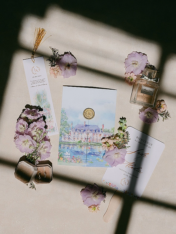 Invitation suite with watercolor wedding invitations, wax seal envelope, rings and lilac flowers on linen backdrop with window light shadows