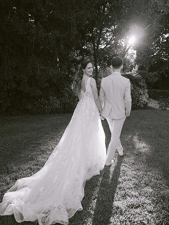 Couple portrait of bride and groom walking away holding hands, bride looking back as her lace train and veil trail in a sunlit garden.