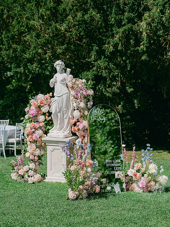 Wedding welcome sign on a clear acrylic arch framed by pastel roses and hydrangeas beside a garden statue on a lawn under tall trees