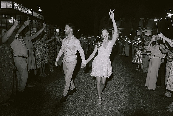 Sparkler exit as newlyweds run hand in hand through a wedding send off tunnel, guests holding sparklers along a gravel path at night