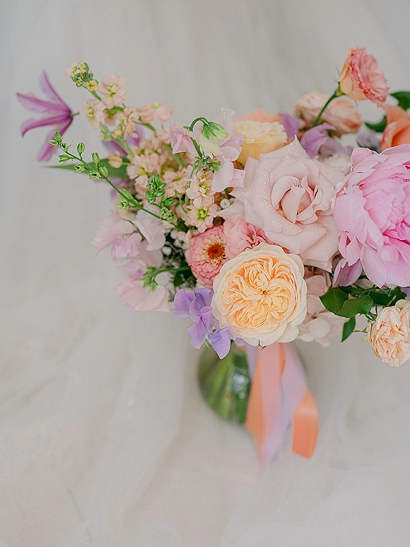 Bridal bouquet of pastel flowers with garden roses, peony, and sweet peas, tied with peach and lavender ribbons in a glass vase on a tabletop