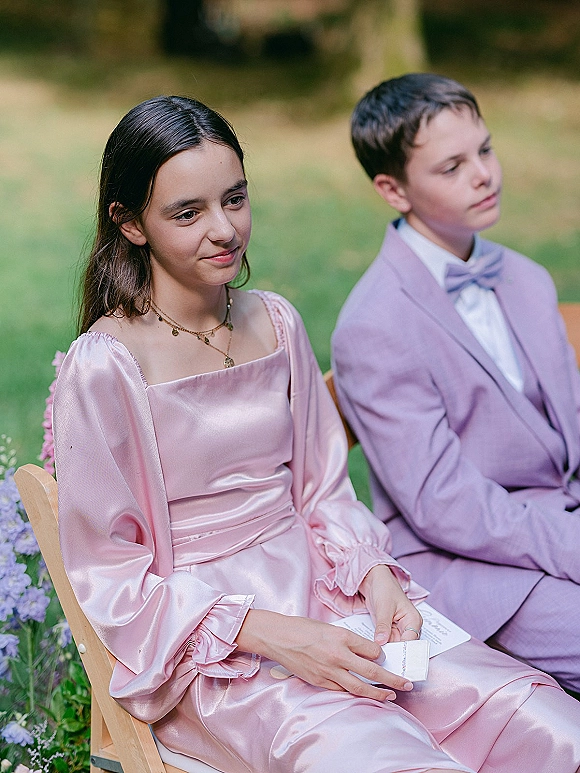Wedding guests watch the ceremony, kids in pink satin dress and lavender suit holding paper programs on wooden chairs on a garden lawn