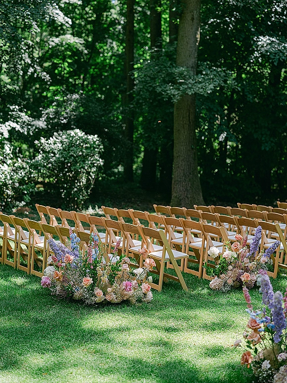 Ceremony seating with outdoor ceremony chairs in neat rows of wood folding chairs, white cushions, and pastel aisle florals on a lawn amid trees