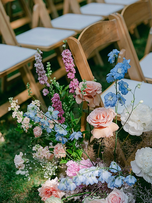 Ceremony aisle flowers in low ground floral arrangements with roses, delphinium, and greenery beside wood folding chairs on a lawn