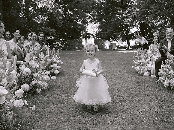 Ring bearer walking down aisle holding a ring pillow, passing floral arrangements and seated guests at an outdoor garden ceremony.