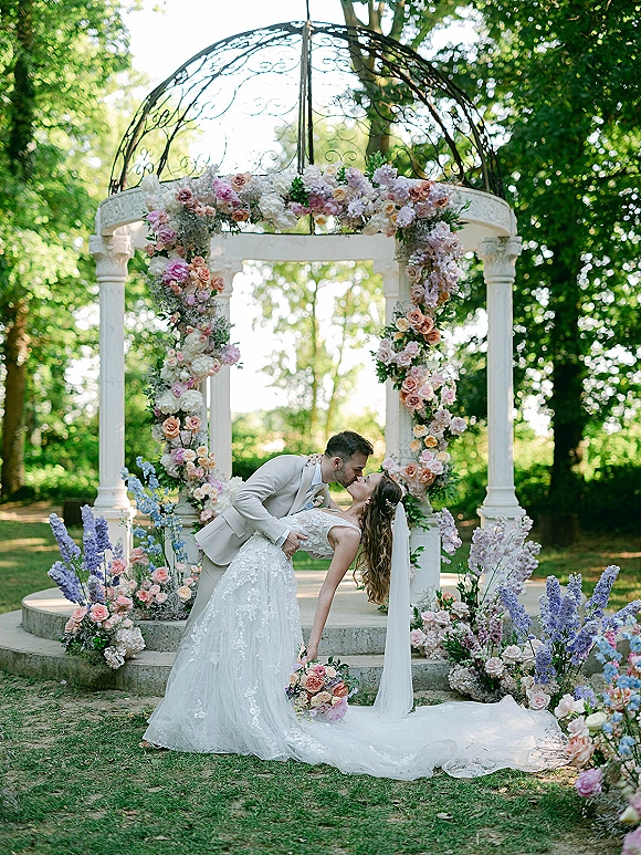 Wedding kiss portrait of bride and groom in a dip kiss beneath a floral gazebo arch, her long veil and lace train flowing in the garden setting