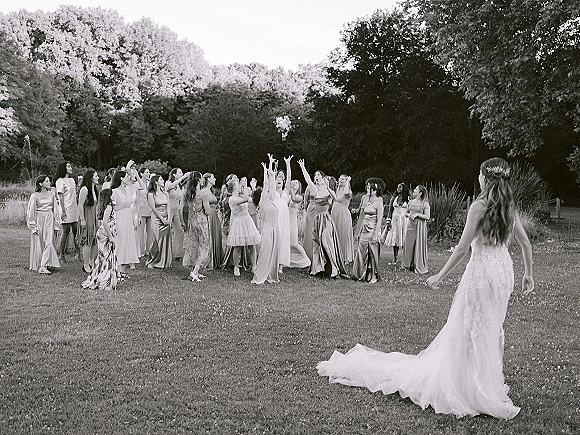 Bouquet toss as the bride tosses a wedding bouquet throw over her veil, bridesmaids in mismatched dresses reaching on a grassy lawn