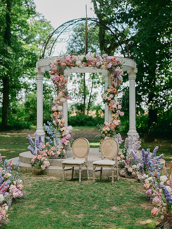 Ceremony altar with outdoor wedding altar floral arch of pastel blooms and greenery, framed by white columns under a wrought iron dome on a garden lawn
