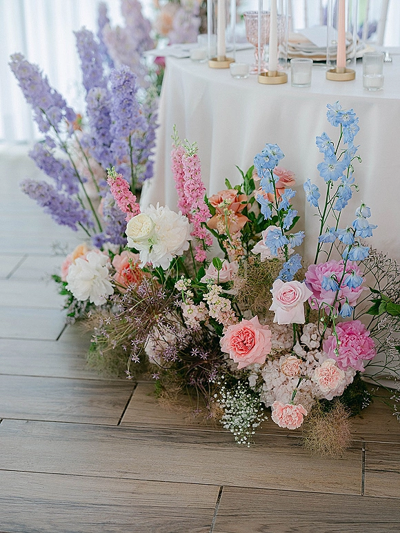 Wedding floral arrangement with pastel florals and tall taper candles in holders along a white-draped reception table with blush goblets