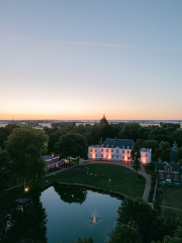 Wedding venue aerial view of an estate wedding venue with manor house, glowing reception tent, and pond fountain at sunset in the countryside