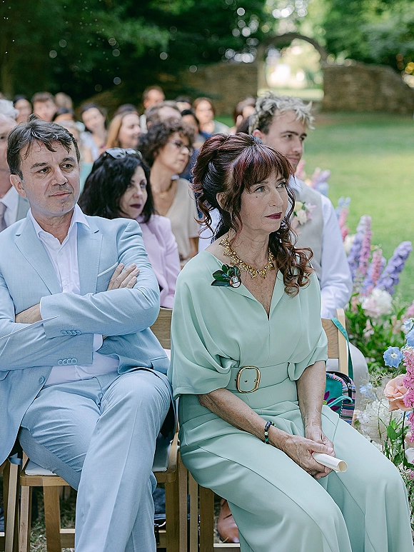 Wedding guests seated in wooden chairs at an outdoor garden ceremony, dressed in pastel attire with corsages and floral aisle arrangements