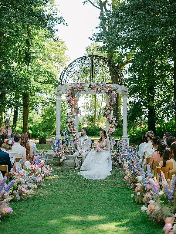 Wedding ceremony moment with bride and groom seated beneath a floral arch at a garden gazebo, pastel aisle flowers and veil in view
