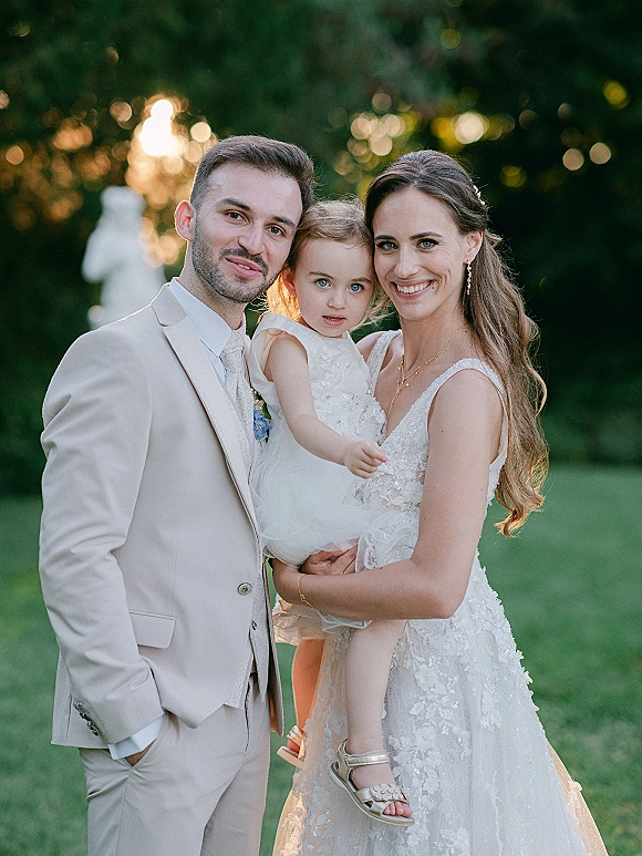 Wedding family portrait of bride and groom with child, bride holding flower girl in white tulle dress on a sunlit garden lawn by a stone statue