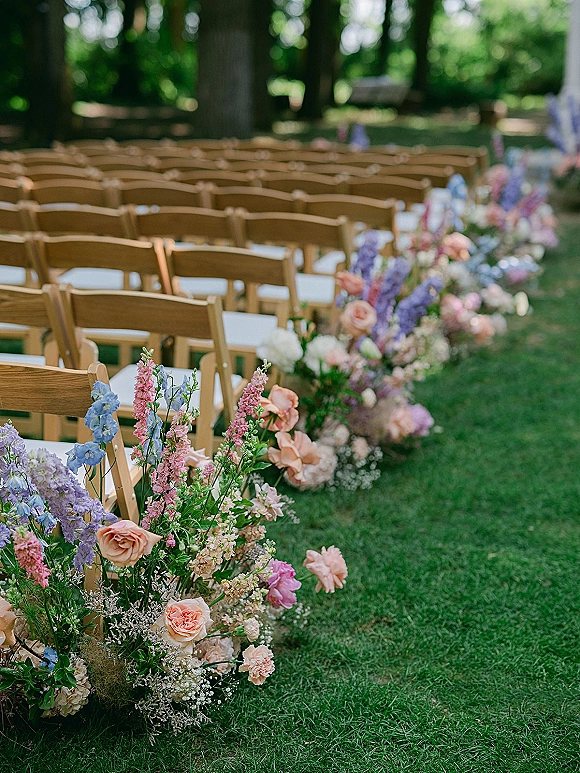 Ceremony aisle decor with wood folding chairs and low rose florals with baby’s breath accents on a garden lawn among trees