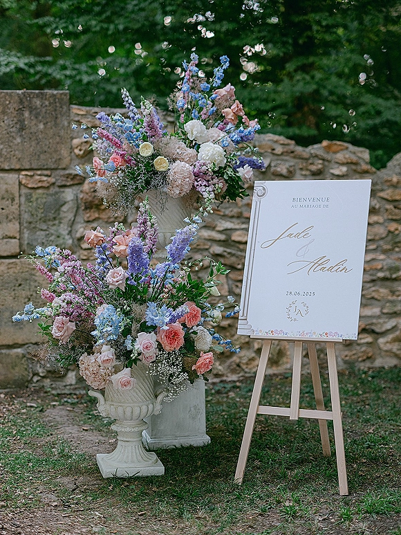 Wedding welcome sign on a wooden easel with roses, delphinium, and baby’s breath in stone urns, set by a stone wall outdoors
