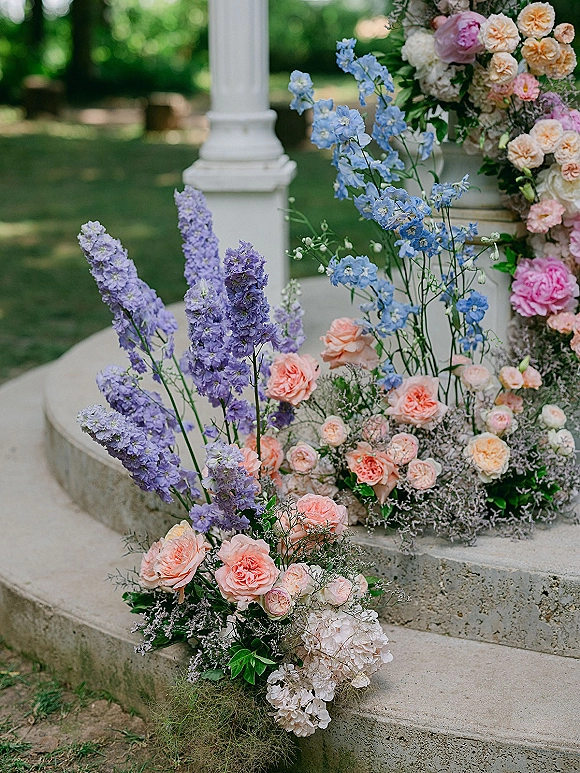 Ceremony florals with pink and peach roses and blue delphinium spilling over stone steps beside a white column on a garden lawn