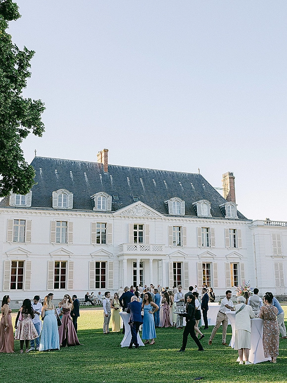 Wedding cocktail hour with guests mingling at outdoor cocktail hour high-top tables with white linens on a lawn by a manor house facade