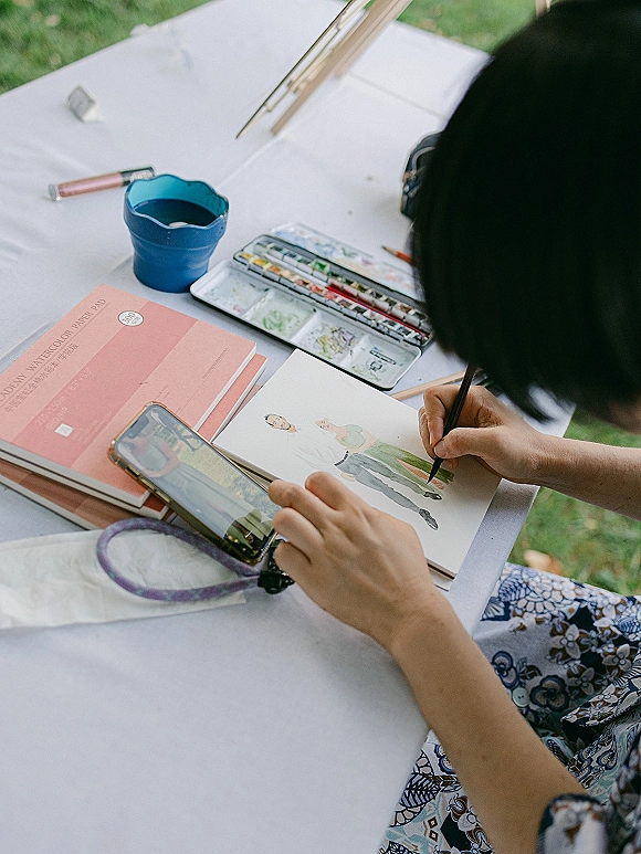 Wedding guest illustration and live wedding painting setup on a white tablecloth, with watercolor palette, brush, sketchbook, and phone on lawn