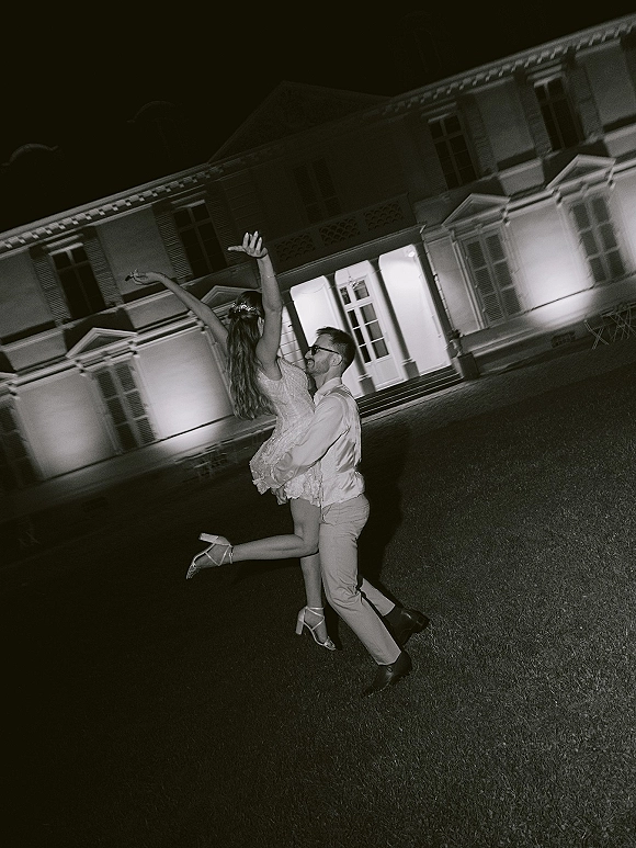 Wedding couple portrait of groom lifting bride in a short wedding dress on a mansion lawn at night, lit windows glowing behind them