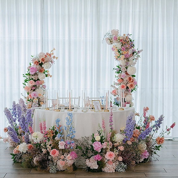 Sweetheart table decor with a round white-clothed table beneath a floral arch, pastel roses and candles against white draping indoors