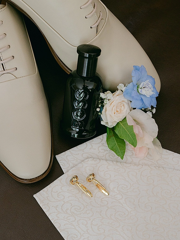 Groom accessories flat lay featuring cream wedding shoes, cologne bottle, boutonniere, gold cufflinks, and pocket square on a dark surface