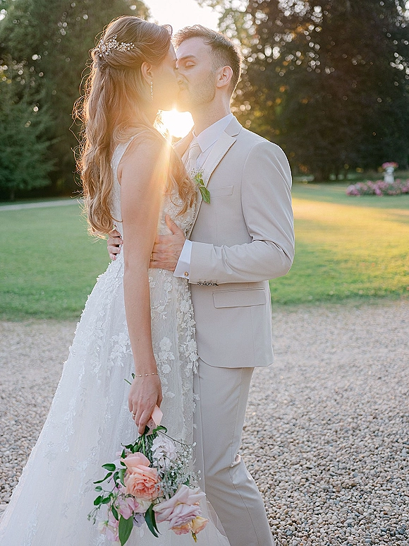 Wedding kiss portrait of bride and groom kissing as she holds a pink rose bouquet, backlit at sunset on a tree-lined lawn with sun flare