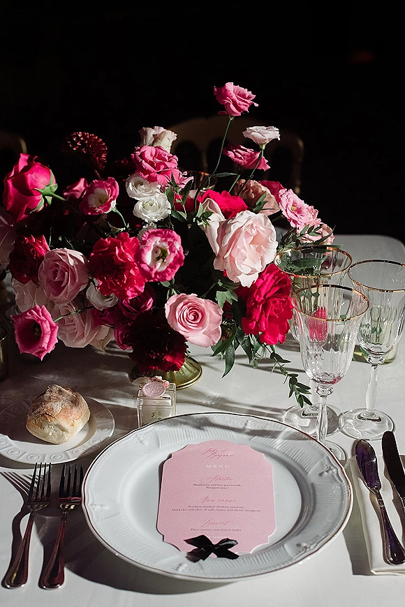 Reception tablescape with wedding table centerpiece of pink roses, black ribbon menu on white plates, gold rim goblets on a white tablecloth