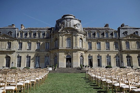Ceremony setup with outdoor ceremony seating of gold chiavari chairs lining a long lawn aisle toward a historic manor facade under blue sky