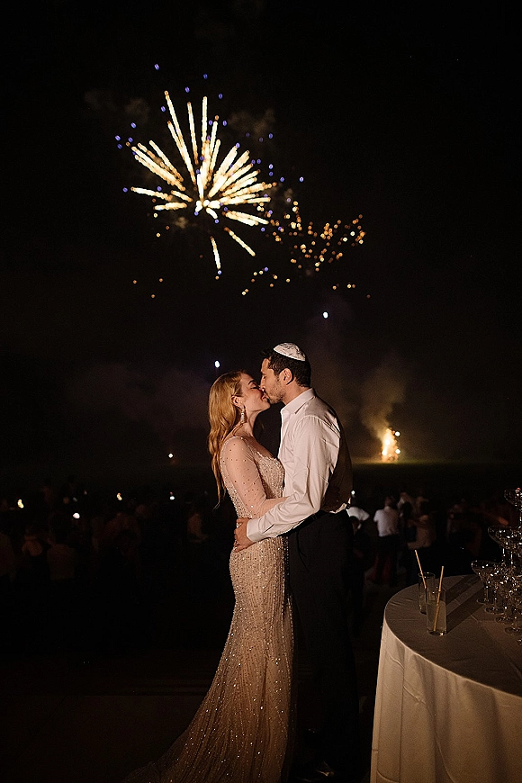 Wedding kiss portrait of newlyweds embracing as fireworks burst overhead, with guests on an outdoor lawn beneath the night sky