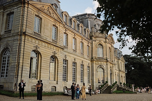 Wedding venue exterior with outdoor lounge seating and cocktail tables in a gravel courtyard before a historic stone building with tall windows