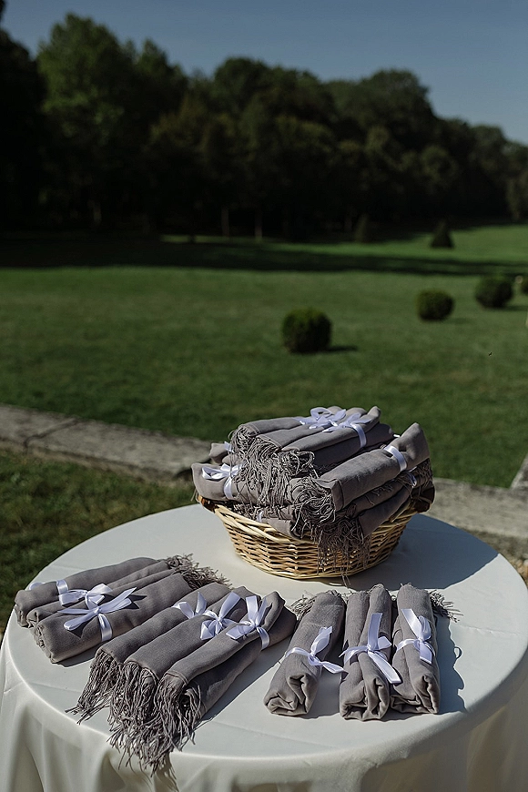 Wedding welcome table with a wedding blanket basket of folded gray blankets tied with white ribbon bows on a lawn by trees and a stone path