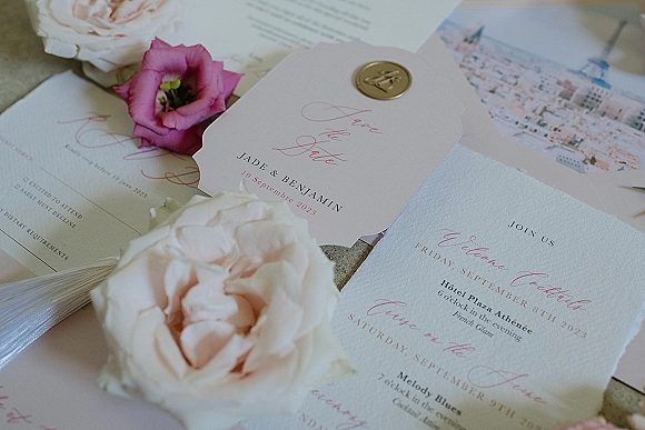 Wedding invitations with calligraphy and deckled edge cards, a wax seal on the envelope, and a blush rose on a stone surface flat lay