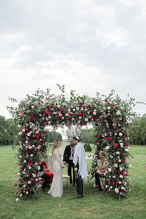 Wedding vows at an outdoor wedding ceremony as bride and groom stand under a rose floral arch with chandelier on a garden lawn