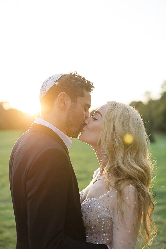 Wedding kiss as newlyweds kissing in profile at sunset, bride in veil and gown with jewelry, groom in black suit and kippah in a green field