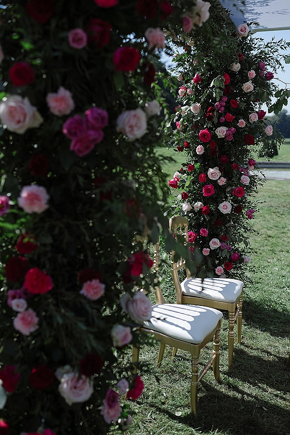 Ceremony floral arch with rose wedding arch pillars draped in greenery garland, framing a lawn aisle with gold chairs under open sky