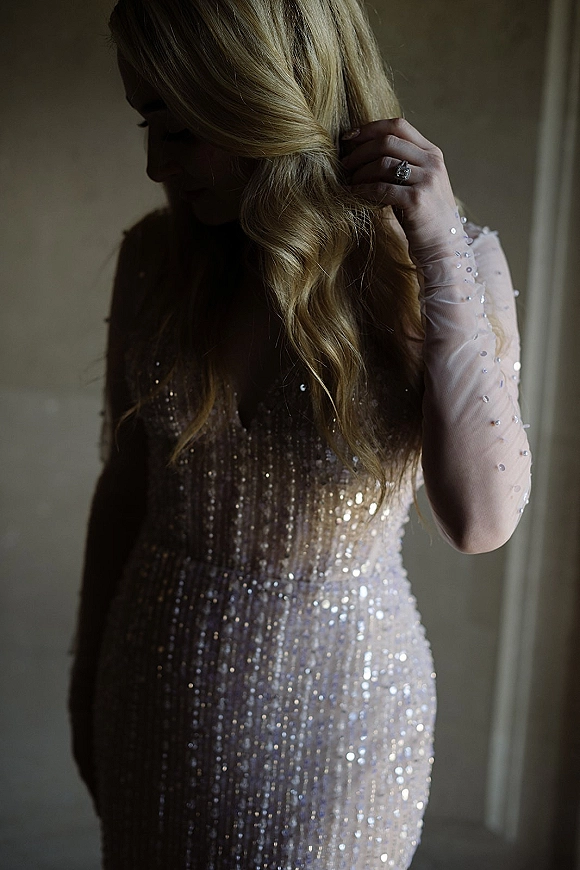 Bridal portrait in a sparkly wedding dress, bride looking down while adjusting her hair, window light highlighting beading and ring