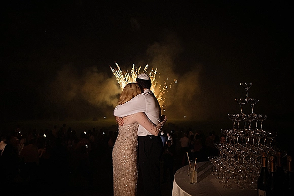 Wedding fireworks moment as newlyweds watching fireworks beside a champagne tower of coupe glasses, guests behind in smoky night sky