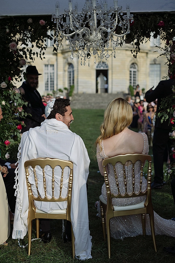 Ceremony moment at a Jewish wedding ceremony as bride and groom sit under a floral chuppah with chandelier on an outdoor lawn with guests